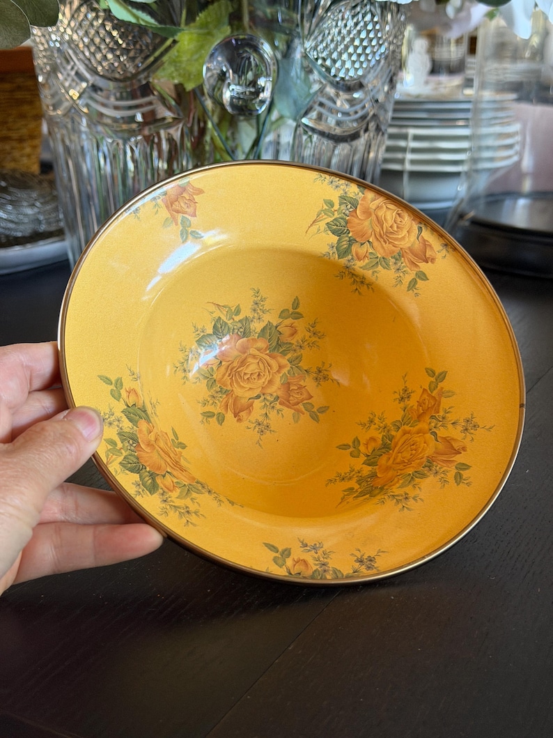 May include: A round, shallow bowl with a golden-yellow base color, featuring a repeating floral pattern of orange and yellow roses with green leaves. The bowl has a thin, metallic gold-toned rim that encircles the entire outer edge of the piece.