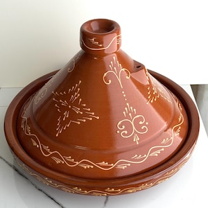 May include: A brown ceramic tagine pot with a conical lid and a round base. The pot is decorated with cream-coloured floral patterns. The tagine is designed for cooking and serving Moroccan and North African dishes.