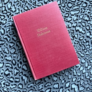 May include: A red hardcover book titled "William Shakespeare" in gold lettering. The book is resting on a patterned surface with a leopard print design in shades of gray and white.