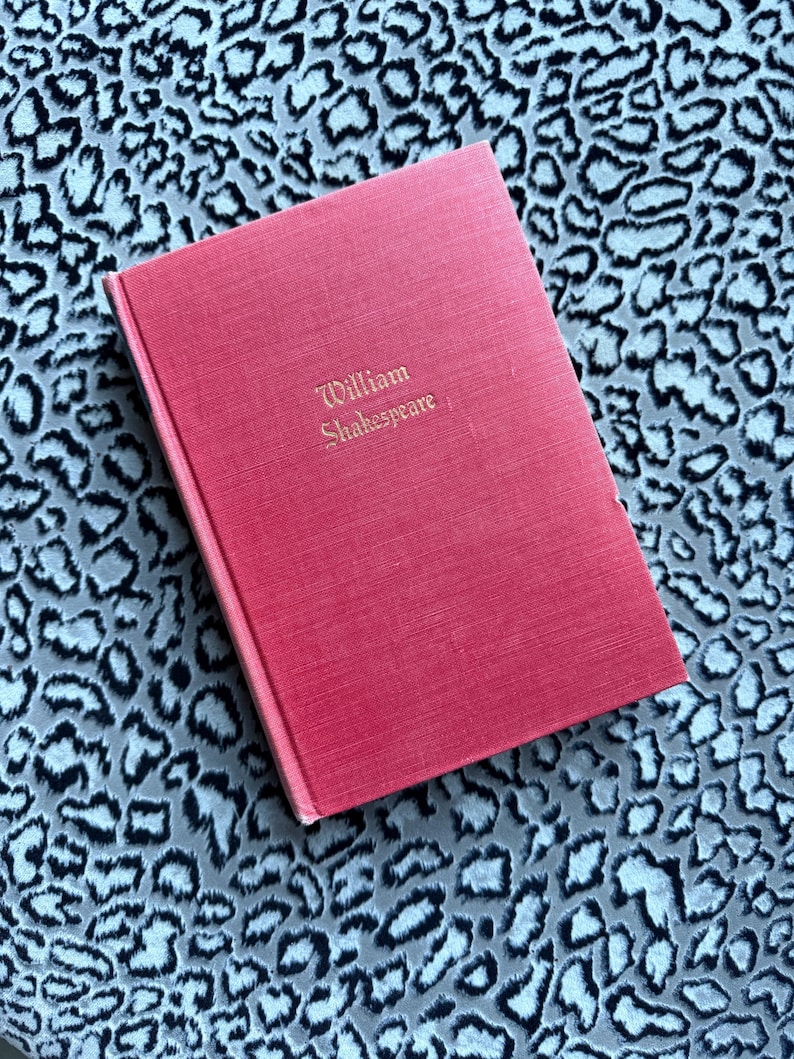 May include: A red hardcover book titled "William Shakespeare" in gold lettering. The book is resting on a patterned surface with a leopard print design in shades of gray and white.