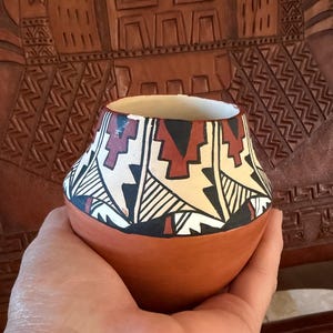 Puede incluir: Una pequeña vasija de barro sostenida en la mano con base redondeada y cuello estrecho. La parte superior presenta un patrón geométrico pintado en negro, rojo oscuro y crema, mientras que la sección inferior es de color marrón terracota.