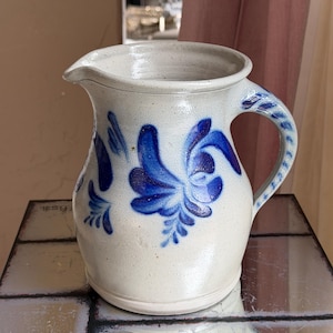 Puede incluir: Una jarra de cerámica gris claro con acabado moteado y un patrón floral pintado a mano en azul. La jarra presenta un pico ancho, un asa curva con detalles decorativos azules y un cuerpo redondeado que se estrecha hacia la base.