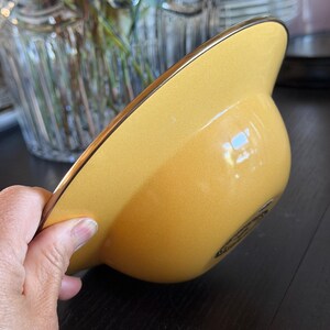 May include: A smooth, solid yellow bowl with a wide, flat rim and a thin metallic gold-toned edge. The bowl features a rounded base and a glossy finish, with a small, dark circular emblem or logo printed on the exterior side near the bottom.