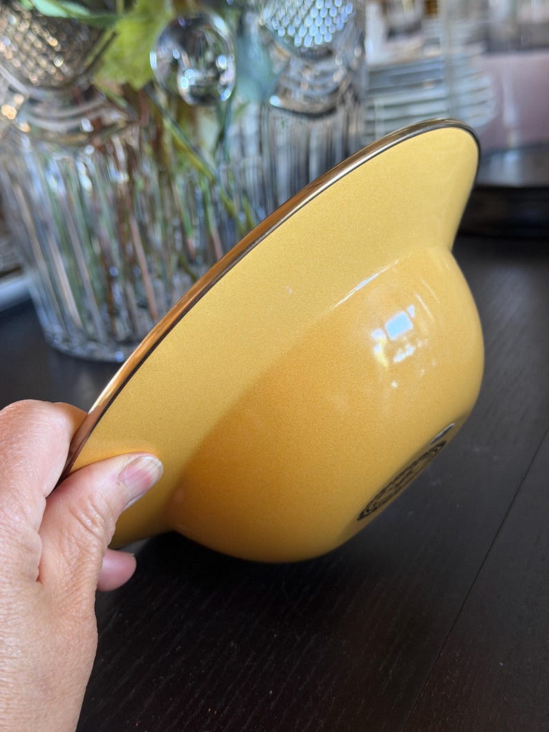 May include: A smooth, solid yellow bowl with a wide, flat rim and a thin metallic gold-toned edge. The bowl features a rounded base and a glossy finish, with a small, dark circular emblem or logo printed on the exterior side near the bottom.