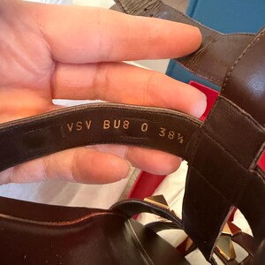 May include: Brown leather sandals with gold-coloured stud accents. The sandals have multiple straps and a buckle closure. The size is indicated as BU8 0 38 1/2.