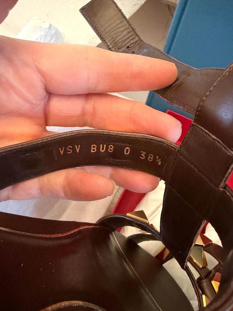 May include: Brown leather sandals with gold-coloured stud accents. The sandals have multiple straps and a buckle closure. The size is indicated as BU8 0 38 1/2.
