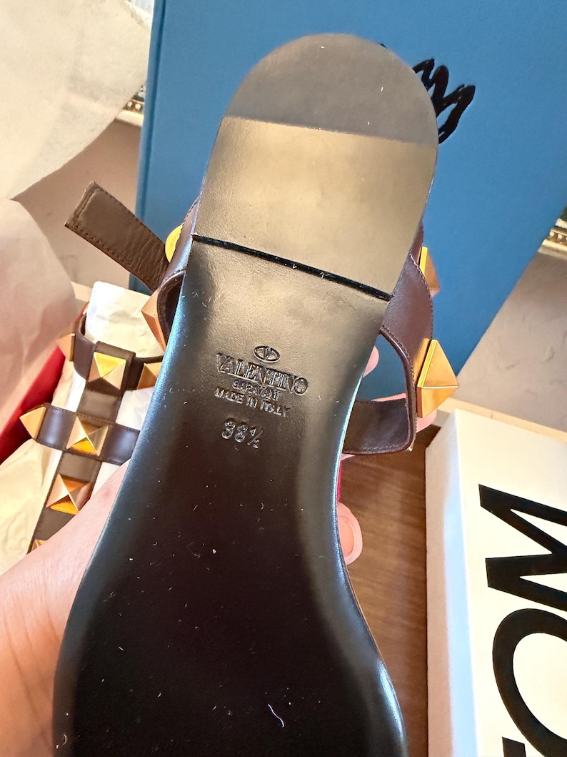 May include: Brown leather sandals with gold pyramid studs. The black sole features the brand name "Valentino" and size "38 1/2". The sandals have a strap and buckle closure. Made in Italy.