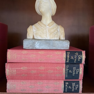 May include: A carved bust of a woman with short hair sits atop a stack of three red books with gold lettering. The books are titled "The Poems of Browning", "The Works of Shakespeare", and "The Works of Balzac". The bust is made of a light-colored stone.