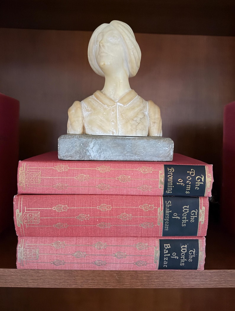 May include: A carved bust of a woman with short hair sits atop a stack of three red books with gold lettering. The books are titled "The Poems of Browning", "The Works of Shakespeare", and "The Works of Balzac". The bust is made of a light-colored stone.
