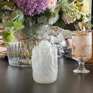 Könnte beinhalten: Ein dekoratives Glasgefäß mit Deckel und geprägtem Blattmuster. Das Gefäß steht auf einer dunklen Oberfläche, im Hintergrund ein Blumenarrangement und andere Glaswaren.