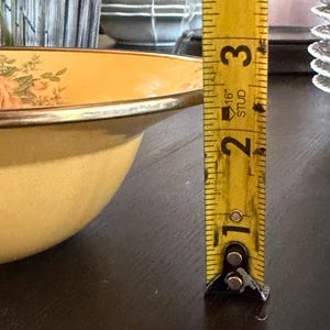 May include: A pale yellow enamel bowl with a floral pattern featuring pink roses and green leaves. A yellow metal measuring tape is positioned vertically next to the bowl, indicating a height of approximately 3.5 inches from the base to the rim.