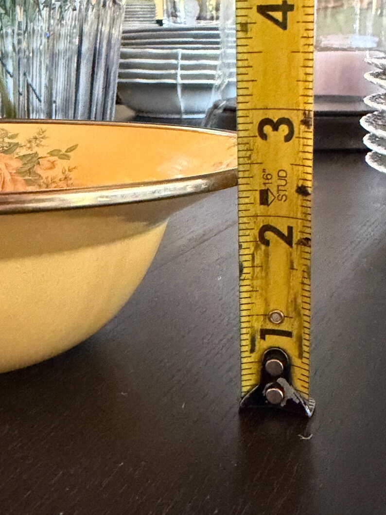 May include: A pale yellow enamel bowl with a floral pattern featuring pink roses and green leaves. A yellow metal measuring tape is positioned vertically next to the bowl, indicating a height of approximately 3.5 inches from the base to the rim.