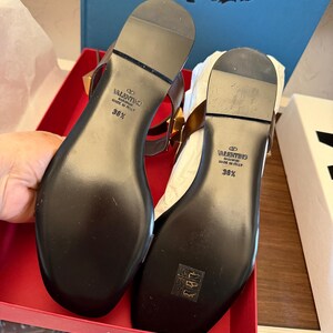 May include: A pair of black leather sandals with gold-tone studs, presented in a red box. The soles are black with the brand name and size 36 1/2. The sandals feature a T-strap design.
