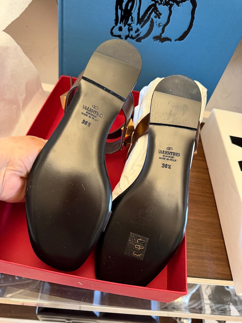May include: A pair of black leather sandals with gold-tone studs, presented in a red box. The soles are black with the brand name and size 36 1/2. The sandals feature a T-strap design.
