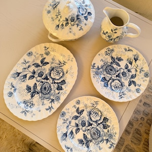 Puede incluir: Un juego de vajilla de porcelana blanca con un diseño floral azul. El juego incluye una fuente con tapa, una jarrita para crema, una fuente ovalada y dos platos redondos. El diseño floral presenta grandes flores y follaje.