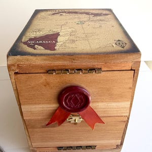 May include: Wooden box with a map of Nicaragua on the lid. The box is rectangular with a hinged lid and a drawer. A red wax seal with a ribbon is on the front. The box is made of light brown wood.