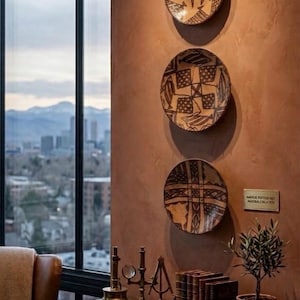 Può includere: Tre piatti circolari in ceramica con motivi geometrici e di pesci marrone scuro montati verticalmente su una parete in terracotta testurizzata. Sotto, una console in legno ospita strumenti scientifici in ottone, una fila di libri e una piccola pianta.