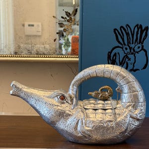 May include: A silver alligator-shaped teapot with an arched handle. The teapot has a textured surface and a small, gold-coloured ram figurine on the lid. The alligator's eye is a red gem.