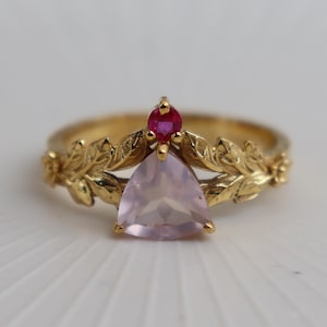 Op de afbeelding: Een gouden ring met een bloemmotief. De ring heeft een driehoekige, lichtroze edelsteen en een kleine, ronde, rode edelsteen. De band is versierd met bladvormige details.