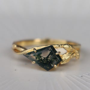 May include: A gold ring featuring a square, dark green gemstone. The ring has a nature-inspired design with leaf accents and a twisted band. The gemstone is set in a gold bezel, displayed on a white surface.