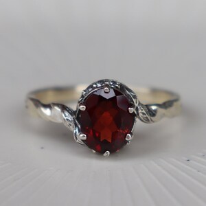 May include: A silver ring featuring a large, oval, deep red gemstone. The band has a twisted design, and the gemstone is set in a decorative bezel. The ring is displayed on a white surface.