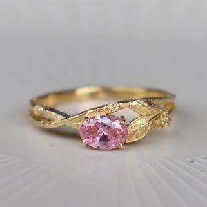 Op de afbeelding: Een gouden ring met een roze edelsteen. De ring heeft een op de natuur geïnspireerd ontwerp met blad- en wijnstokdetails. De ovale roze edelsteen is het middelpunt, gezet in een delicate gouden band. De ring wordt gepresenteerd op een wit oppervlak.