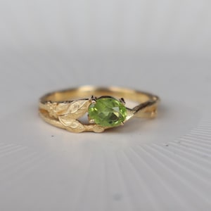 May include: A gold ring featuring an oval-cut, light green gemstone set in the centre. The band is crafted with a textured, matte finish and detailed with sculpted leaf motifs that wrap around the sides of the central stone setting.