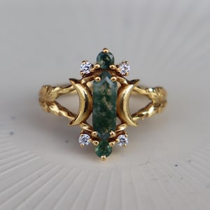 May include: A gold ring featuring a central elongated moss agate stone flanked by two crescent moon shapes. The setting includes four small clear round stones and two small round moss agate accents. The band has a textured leaf-like design.