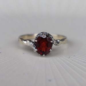 May include: A silver ring featuring a central oval-cut deep red garnet gemstone held in a prong setting. The band of the ring has a twisted rope-like texture that extends from the sides of the stone setting, creating a continuous metallic design.
