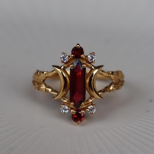 May include: A gold ring featuring a central vertical marquise-cut deep red garnet stone flanked by two crescent moon shapes. The design includes two smaller round red garnet stones at the top and bottom, accented by four small clear white stones.