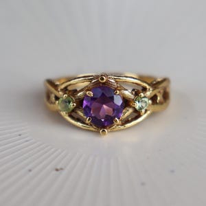 May include: A gold ring featuring a central round-cut purple amethyst gemstone flanked by two smaller round-cut light green peridot gemstones. The band has a textured, organic, branch-like design that wraps around the stones in a cage setting.