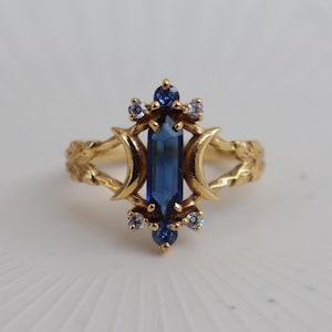 Puede incluir: Un anillo de oro con una piedra preciosa azul hexagonal alargada en el centro, flanqueada por dos formas de media luna. La banda está decorada con pequeñas piedras redondas azules y transparentes, con grabados de hojas en los laterales.