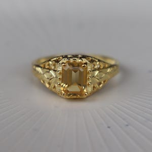 May include: A gold ring featuring a central emerald-cut yellow gemstone. The band displays an intricate openwork design with textured metal detailing surrounding the stone, creating a decorative frame that highlights the faceted yellow gem.