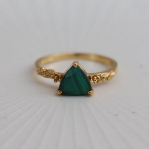 Peut inclure: Une bague en or ornée d'une pierre précieuse en malachite triangulaire au centre. L'anneau est façonné avec un motif en relief détaillé représentant de petites feuilles et des fleurs s'étendant de chaque côté du sertissage de la pierre.