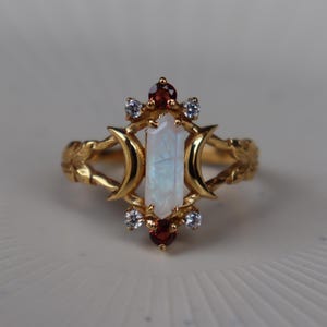 Op de afbeelding: Een gouden ring met een centrale zeshoekige iriserende maansteen, geflankeerd door twee halvemaanvormen. De zetting bevat twee kleine ronde rode granaatstenen aan de boven- en onderkant, geaccentueerd door vier kleine heldere witte steentjes.