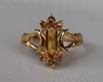 Puede incluir: Un anillo de oro con una piedra preciosa amarilla central de corte baguette vertical flanqueada por dos formas de media luna. La banda está decorada con patrones de hojas y pequeñas piedras amarillas redondas engastadas en la parte superior e inferior.