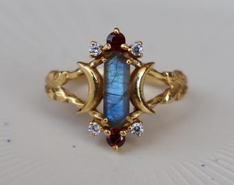 May include: A gold ring featuring a central rectangular labradorite stone flanked by two crescent moon shapes. The design includes four small clear round stones and two small round red stones positioned at the top and bottom of the centre.