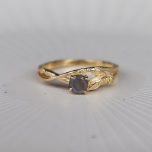 Può includere: Un anello in oro con una pietra preziosa rotonda dai riflessi blu-grigi, incastonata in una fascia che ricorda rami intrecciati con piccole foglie testurizzate. Il metallo ha una finitura lucida e la pietra è fissata da quattro piccole griffe.