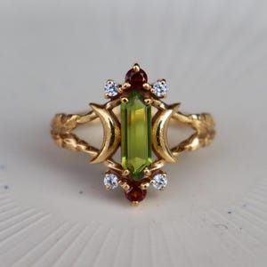 Op de afbeelding: Een gouden ring met een centrale groen peridoot in baguette-slijpvorm, geflankeerd door twee halve maanvormen. De zetting bevat twee kleine ronde rode granaatstenen aan de boven- en onderkant, geaccentueerd door vier kleine heldere witte stenen.