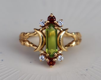 May include: A gold ring featuring a central baguette-cut green peridot stone flanked by two crescent moon shapes. The setting includes two small round red garnet stones at the top and bottom, accented by four small clear white round-cut stones.