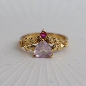 Op de afbeelding: Een gouden ring met een grote, lichtroze, in trillion-geslepen edelsteen in het midden, bekroond met een kleine, ronde, felrode edelsteen. De ringband is voorzien van een gedetailleerd, verhoogd patroon van bladeren en ranken.