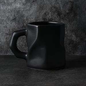 May include: A matte black ceramic mug with a unique, textured design. The mug has a square base and a rounded handle. The exterior has an uneven, sculpted appearance. The mug is designed for hot or cold beverages.