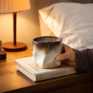 Könnte beinhalten: Eine handgefertigte Keramik-Tasse mit einem einzigartigen, strukturierten Design. Die Tasse hat einen Farbverlauf, der von einem dunkelbraunen Rand zu einer hellbeigen Basis übergeht. Sie steht auf einem Buch mit dem Wort "FLOWERS".