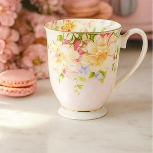 May include: A white ceramic mug with a floral design in yellow, pink, and purple. The mug has a handle and a gold rim. The mug is on a white surface. Two pink macarons are in the background. The mug is likely for tea or coffee.