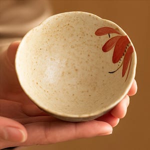 May include: A small, off-white ceramic bowl with a speckled texture and a scalloped rim. The bowl features a hand-painted red floral design on the interior. The bowl is held in a person's hand, showcasing its size and shape.