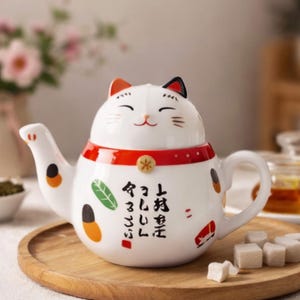 May include: White ceramic teapot with a cat face design. The teapot features a red collar, orange and black accents, and Japanese characters. It sits on a wooden tray with sugar cubes.