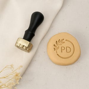 May include: A wax seal stamp with a black handle and gold-colored stamp head, next to a tan wax seal with a leaf design and the letters "PD" inside a circle. The stamp and seal are on a white surface.