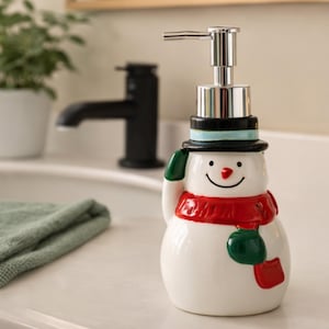 以下が含まれることがあります： 銀色のポンプが付いた、陶器製の雪だるまのソープディスペンサー。雪だるまは黒いシルクハット、赤いマフラー、緑色のミトンをしています。ディスペンサーは白く、笑顔の顔が描かれています。