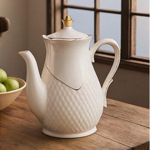 May include: A cream-coloured ceramic teapot with gold-coloured detailing. The teapot features a quilted pattern and a curved handle. It is placed on a wooden surface.