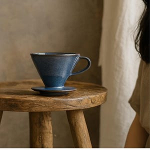 Könnte beinhalten: Ein blauer Keramik-Kaffee-Aufgießer mit passender Untertasse. Der Aufgießer hat eine konische Form mit einem Griff und einem dunklen Rand. Er steht auf einem kleinen, runden Holzschemel.